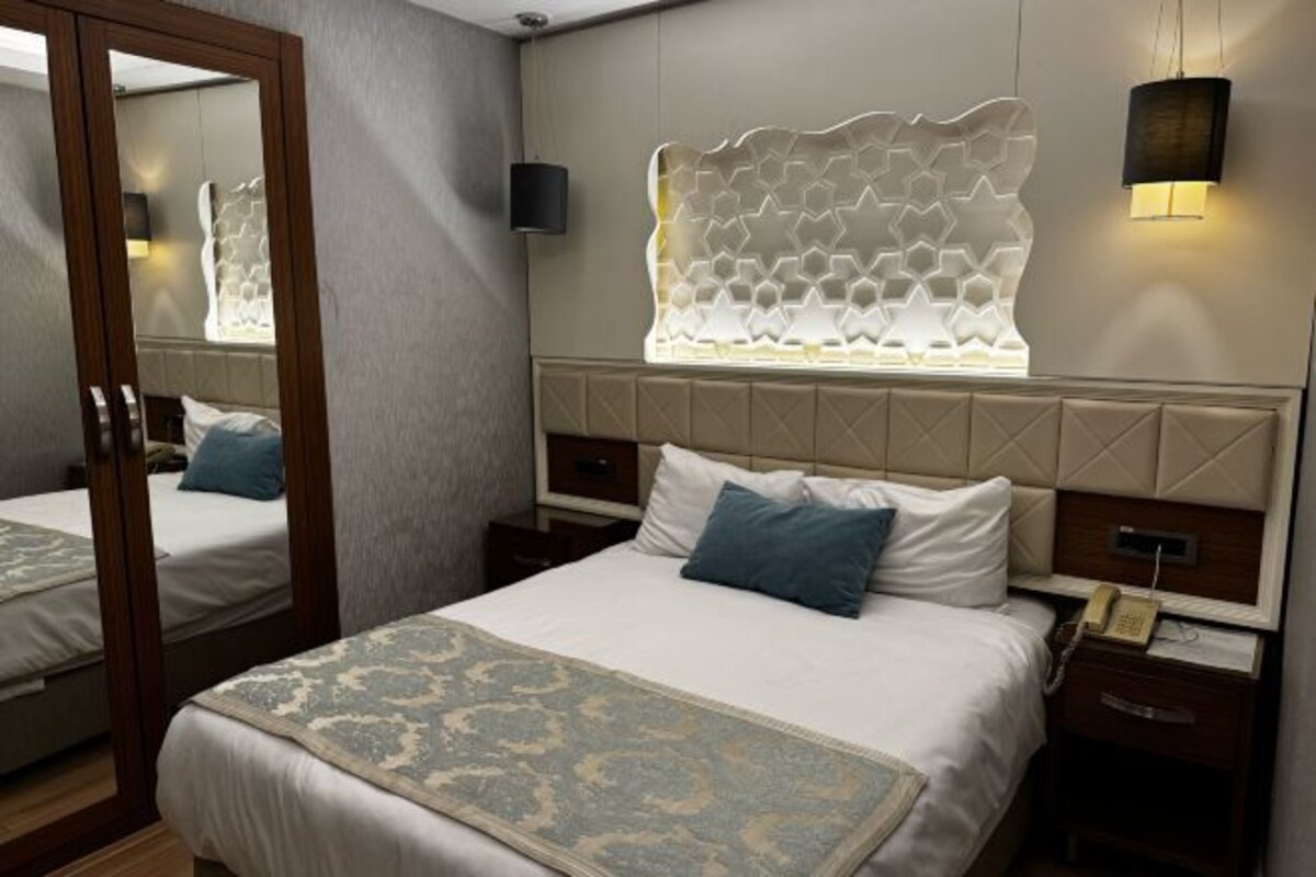 A hotel room with a large bed, two bedside tables, a mirror, a decorative wall panel, and a wall-mounted lamp.