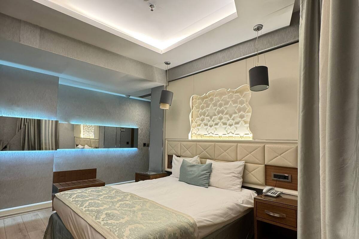 A modern hotel room with a large bed, a bedside table with a lamp, a headboard with a geometric light design, and a ceiling light fixture.