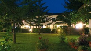 Une cour d’hôtel bien éclairée la nuit, avec un grand palmier, des plantes en pot et un bâtiment doté de nombreux balcons.