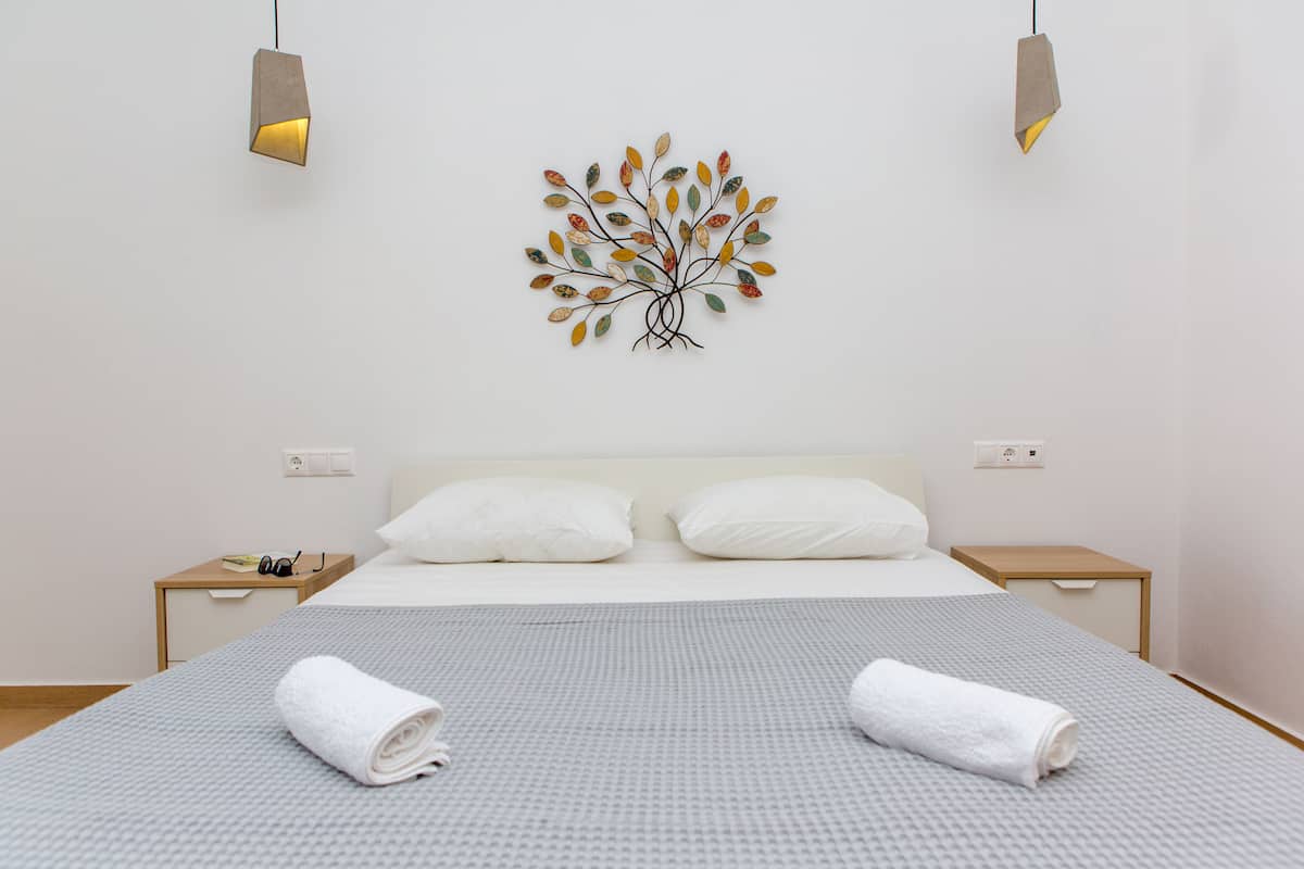 Cama bem arrumada com um edredom cinza texturizado, dois travesseiros brancos e duas toalhas brancas enroladas. Há uma obra de arte na parede, representando uma árvore com folhas multicoloridas e tronco marrom. Duas mesas de cabeceira com abajures estão de cada lado da cama.