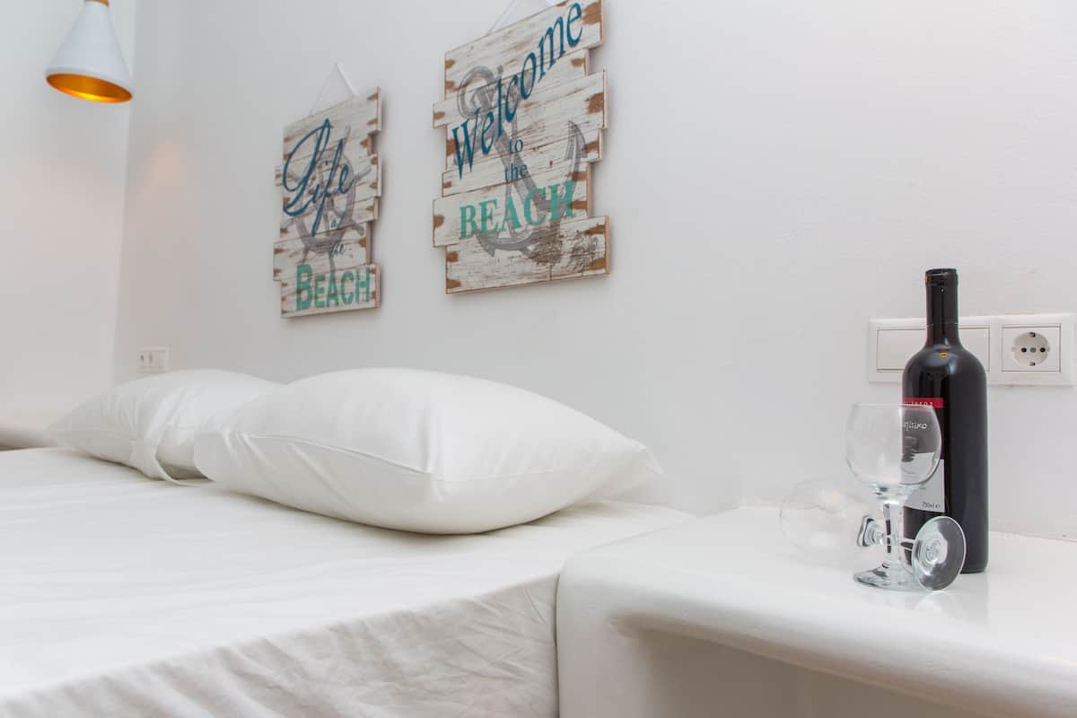 Cama bem arrumada com roupa de cama e almofadas brancas, uma garrafa de vinho e um copo em uma mesa de cabeceira, e dois sinais de madeira na parede com a inscrição 'Welcome to the Beach'.