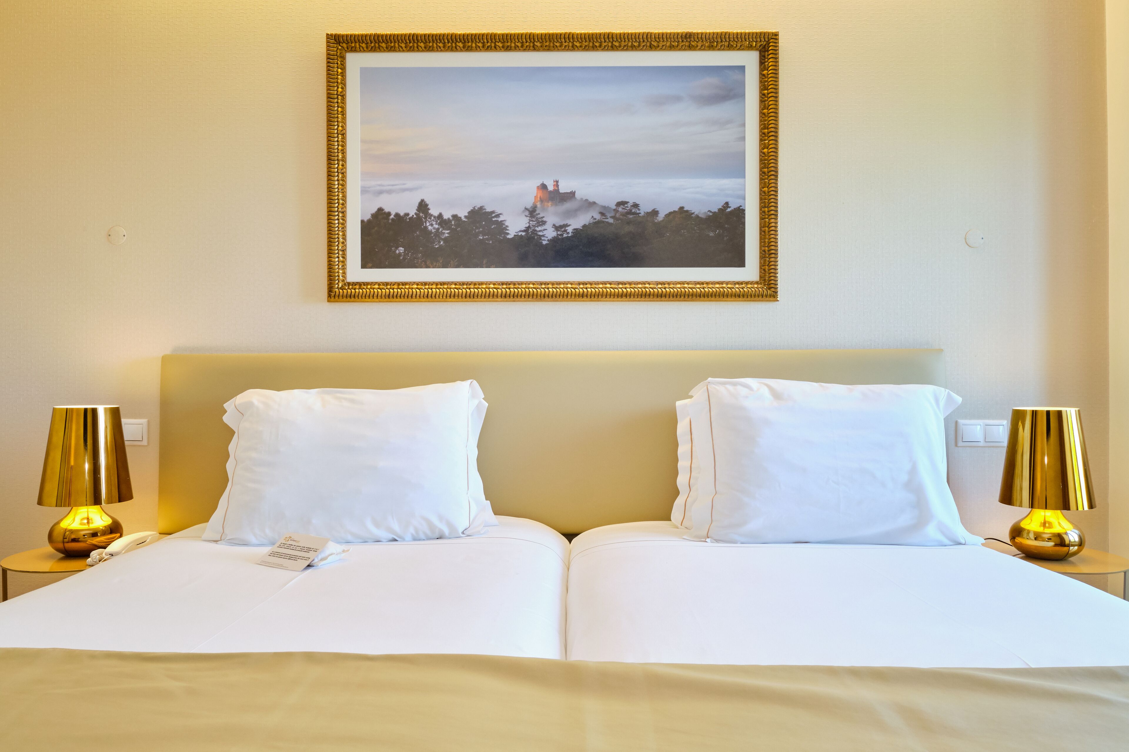 Uma cama de casal com roupa de cama branca, duas luminárias de cabeceira e um quadro na parede.