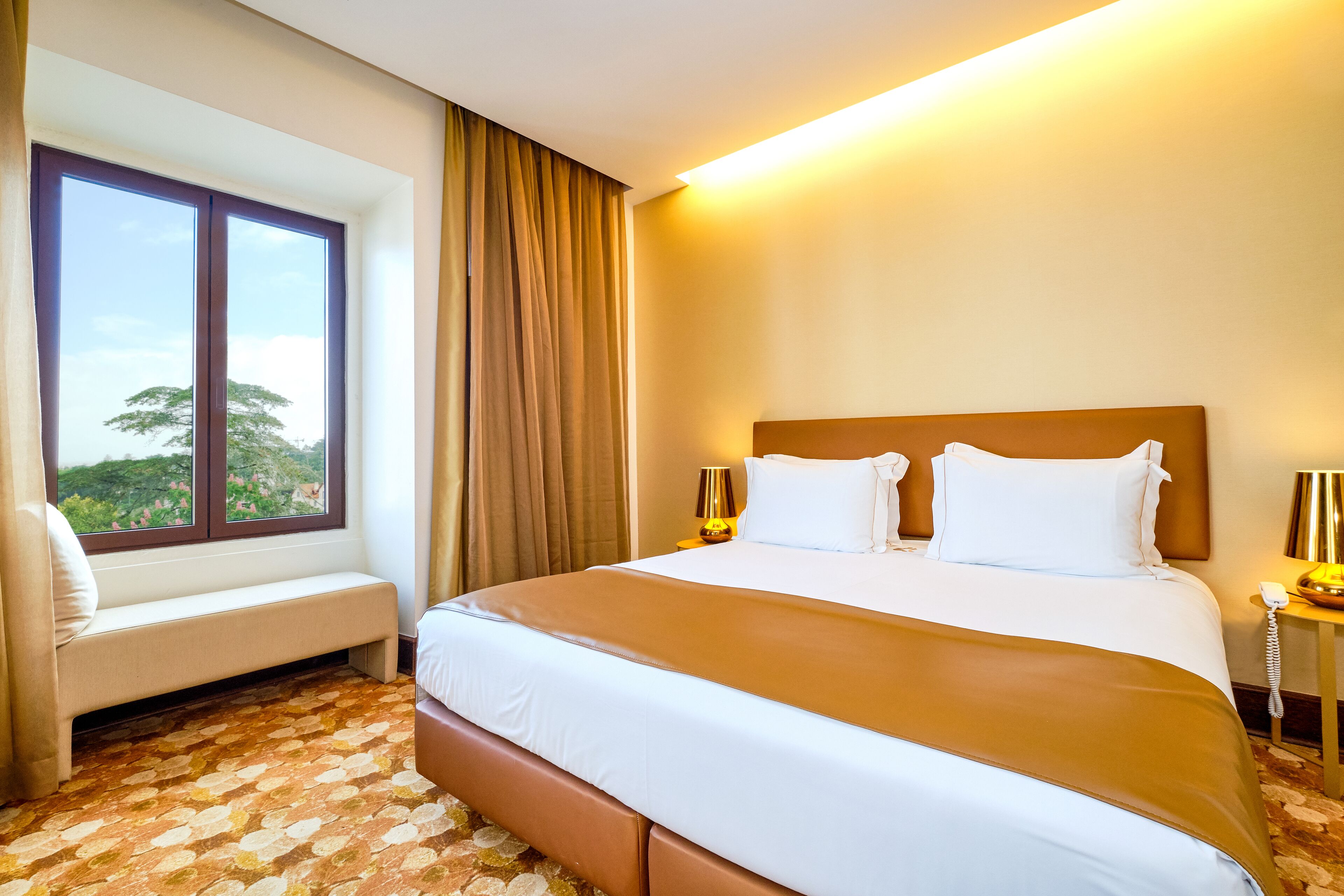 Hotellihuone, jossa on suuri sänky, ikkunan vieressä oleva penkki ja näkymä vehreään maisemaan.