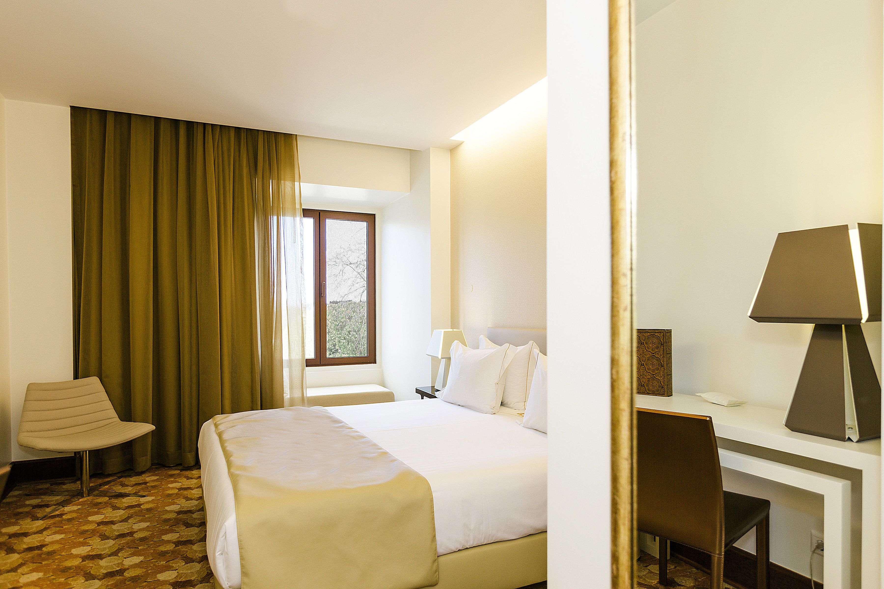 Quarto de hotel com cama, escrivaninha, cadeira, abajur e janela com cortinas.