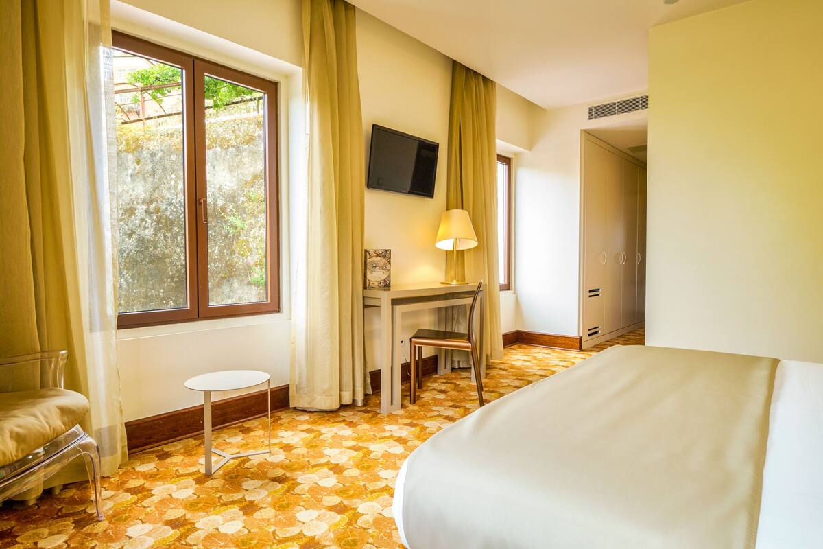 A hotel room with a bed, a desk with a chair, a TV mounted on the wall, and a window with curtains.