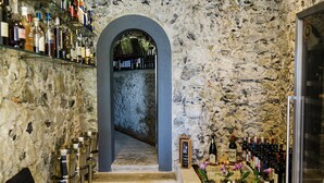 Zona de bar con paredes de piedra, varios botellas y vasos, una escalera que baja a un nivel inferior, y una exhibición de flores.