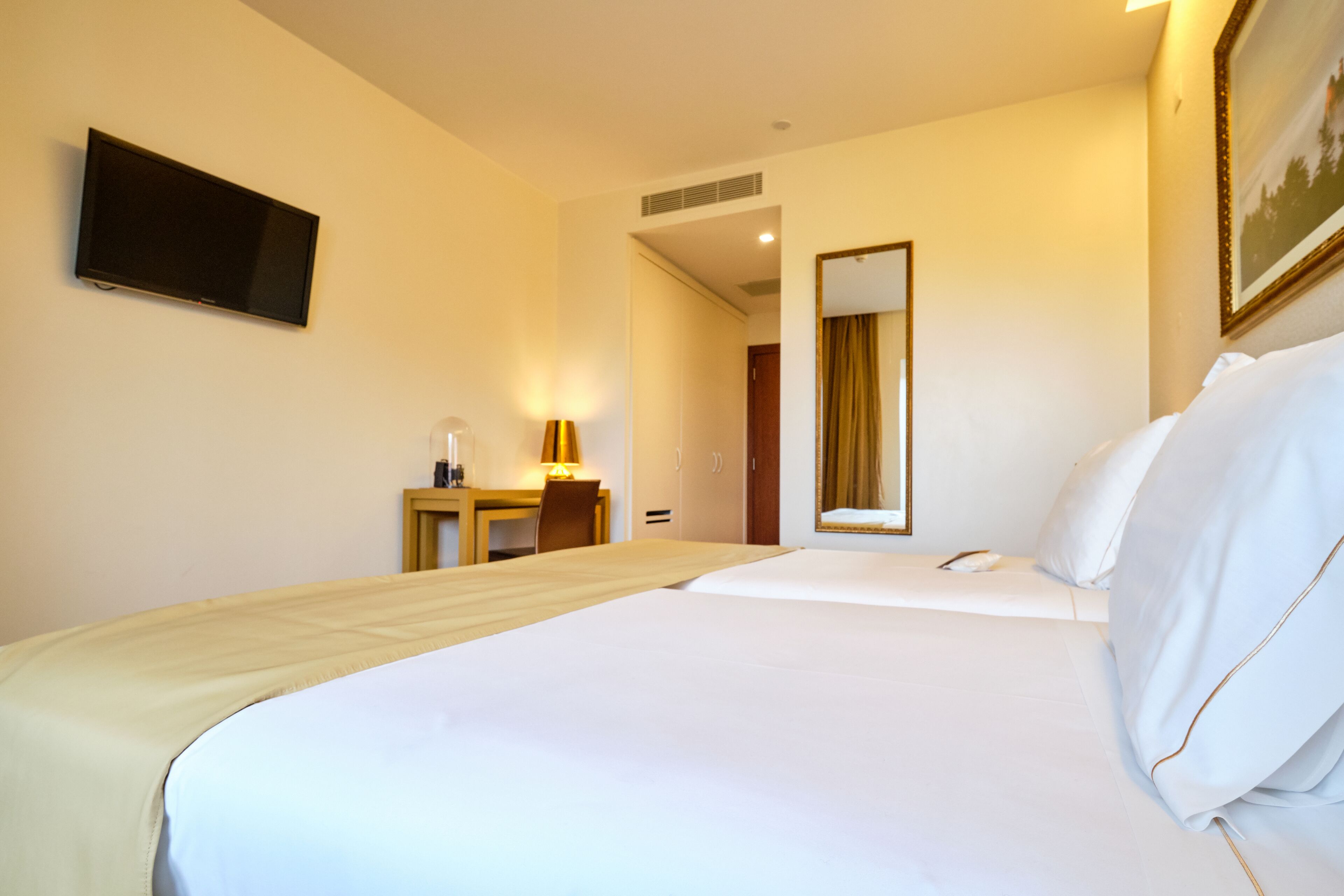 Una habitación de hotel con una cama, un escritorio con lámpara, un televisor y un espejo.