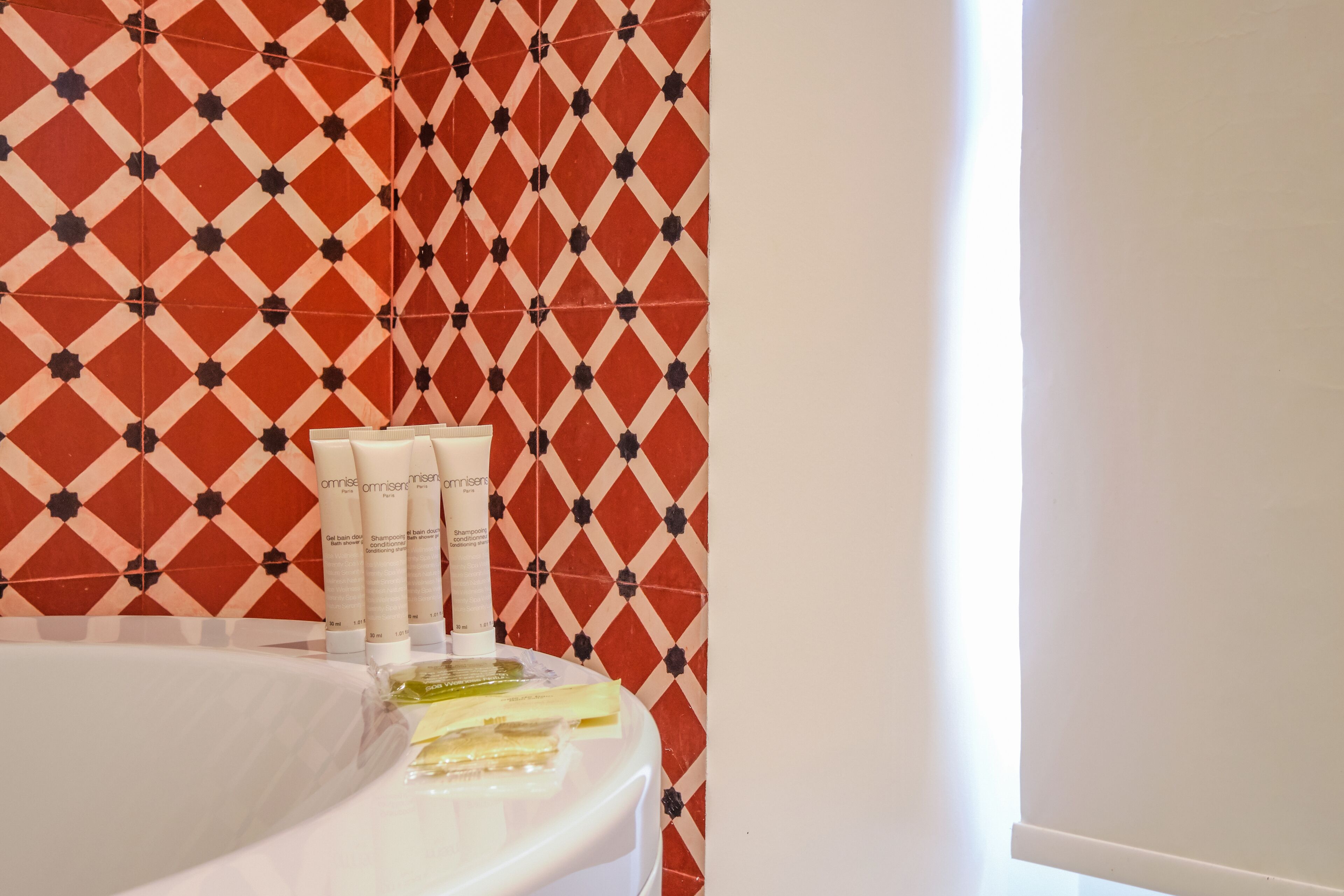 A bathroom with a patterned tile wall, a white bathtub, and toiletries on the edge.
