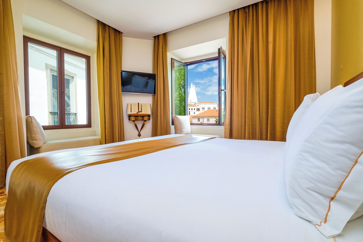 Een hotelkamer met een groot bed, een flatscreen televisie aan de wand, een raam met gordijnen en uitzicht op een gebouw met een toren.