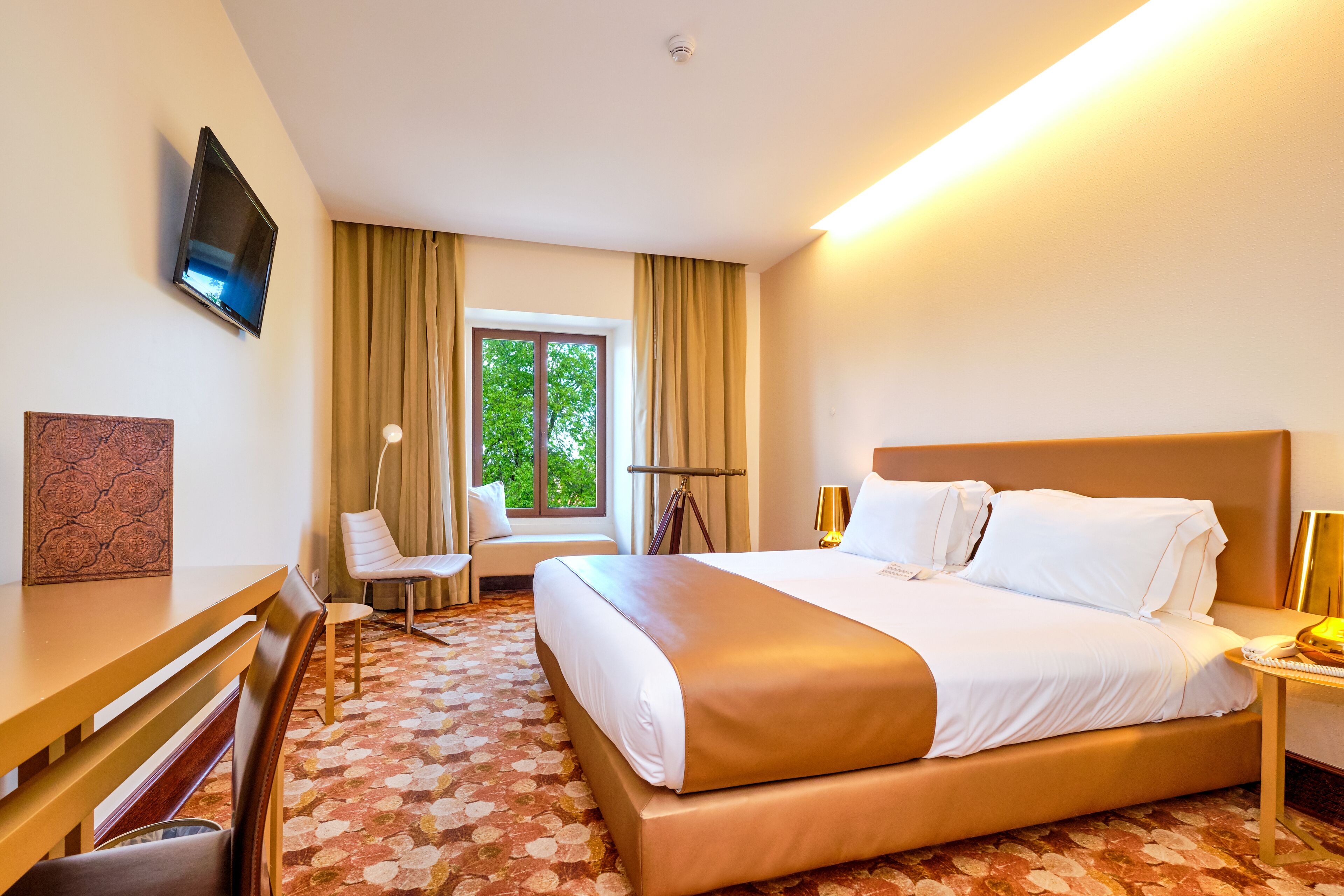 A hotel room with a large bed, a desk, a chair, a TV mounted on the wall, and a window with curtains.