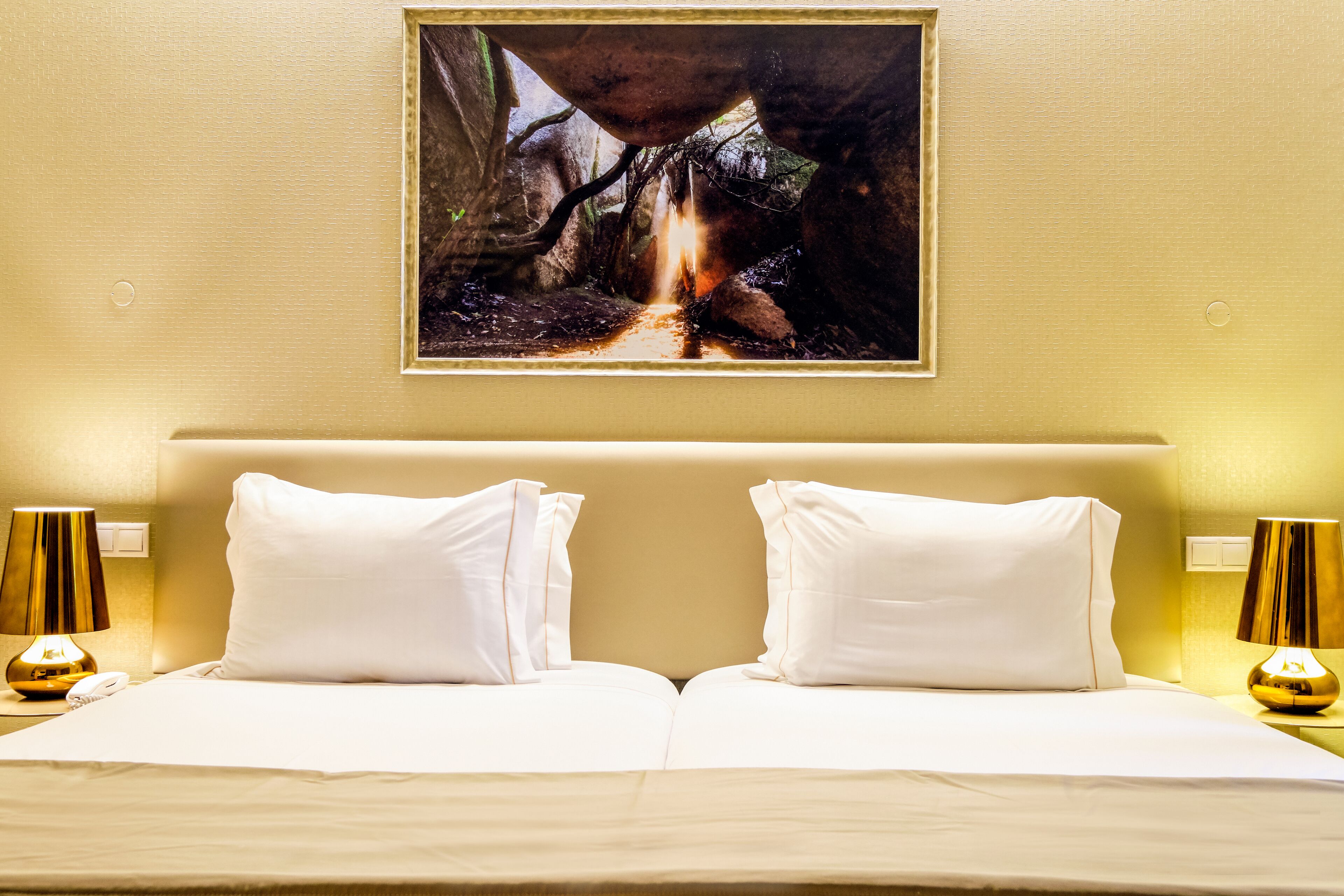 A hotel room with a large bed, two white pillows, a painting of a cave, and two lamps on either side of the bed.