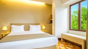 Habitación de hotel con una cama grande, lámparas de noche, un banco y una ventana con vistas a zonas verdes.