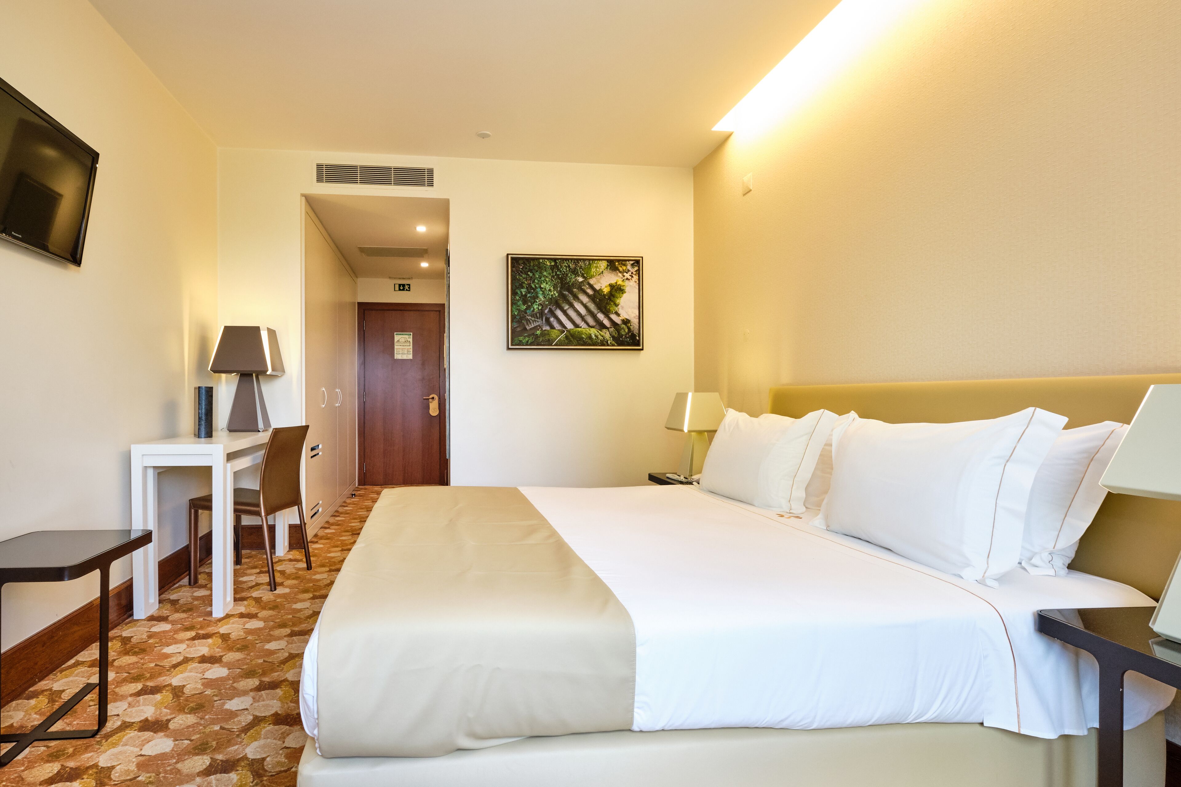A hotel room with a large bed, a desk, a chair, a lamp, and a painting on the wall.