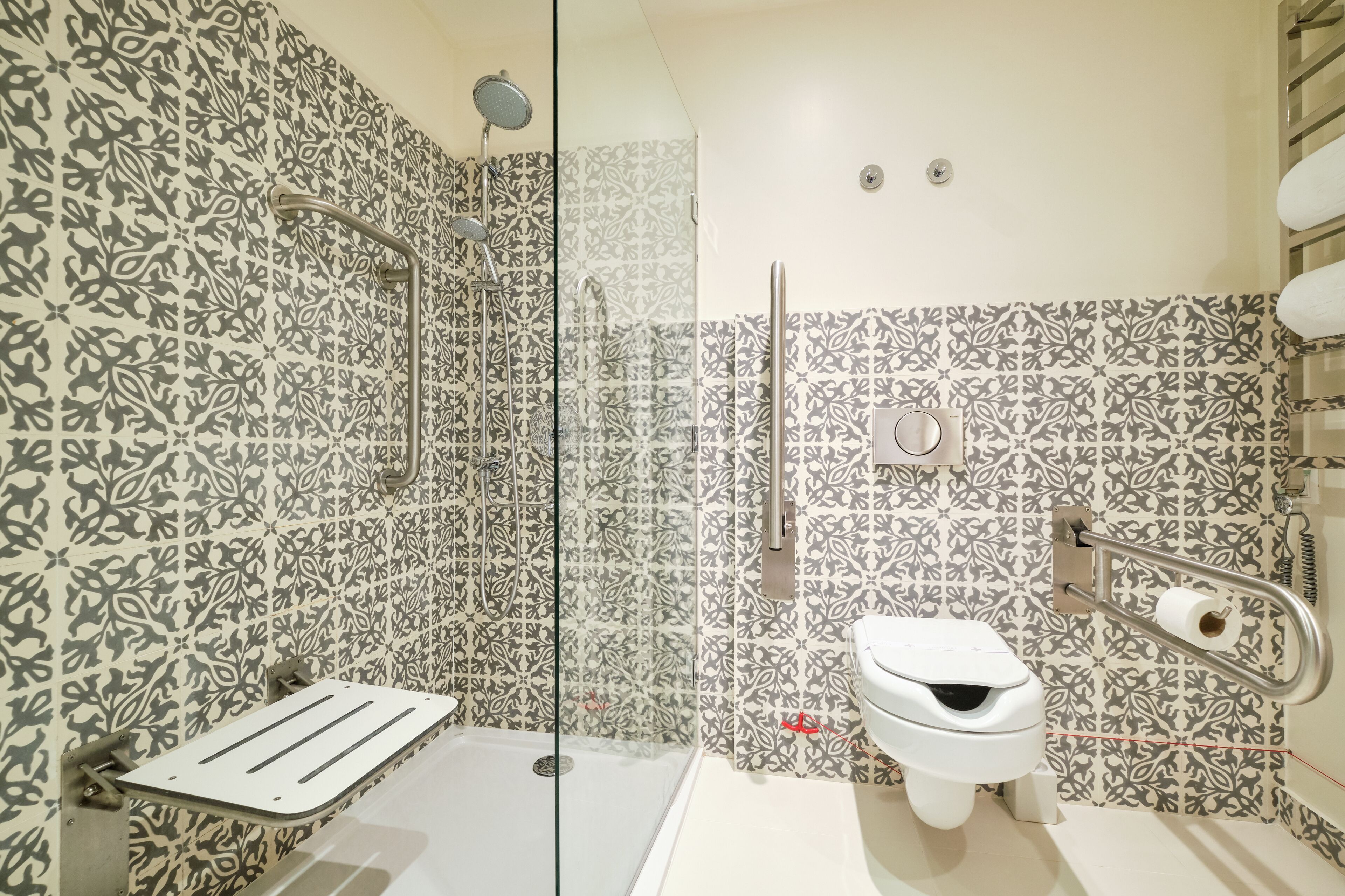 Une salle de bain avec des carreaux décorés, une douche en verre, des toilettes blanches et une barre d’appui.