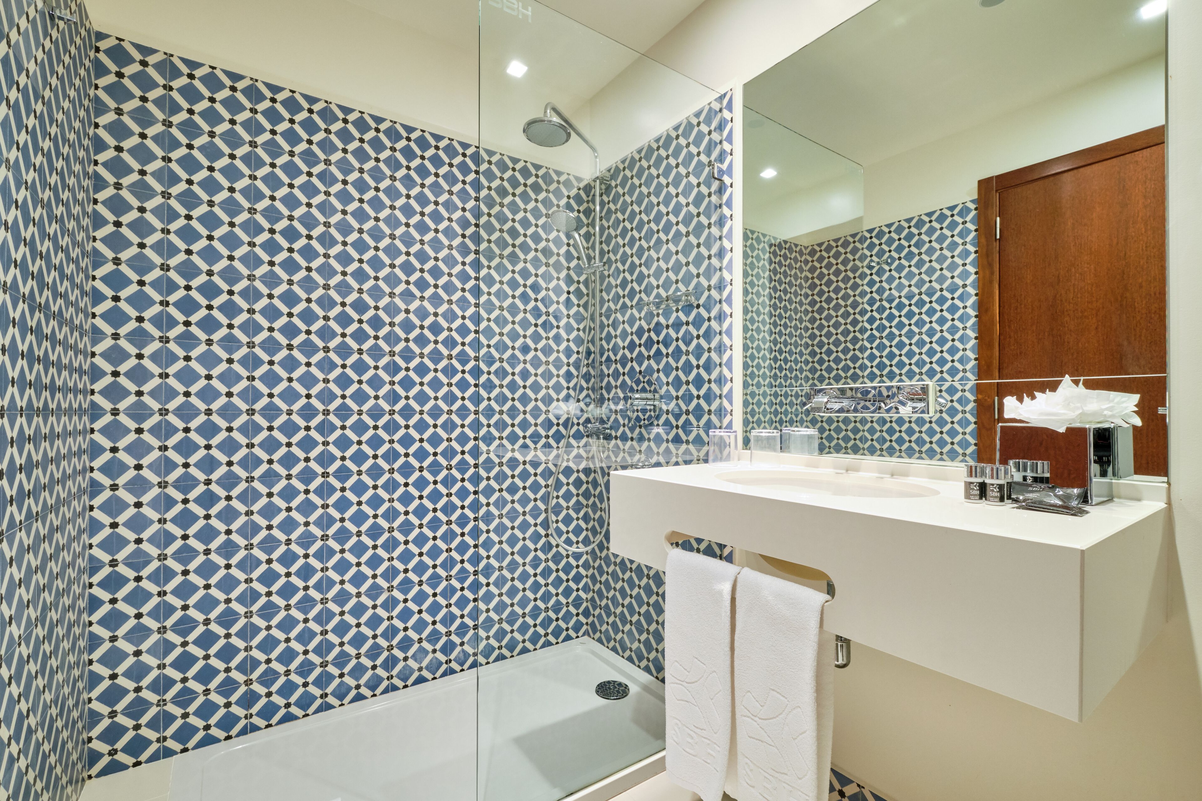 Une salle de bain moderne avec des carreaux bleus et blancs à motifs, une douche en verre, un lavabo blanc avec des articles de toilette et un meuble en bois.