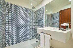 Un baño moderno con baldosas azules y blancas estampadas, una mampara de cristal, un lavabo blanco con artículos de aseo y un armario de madera.