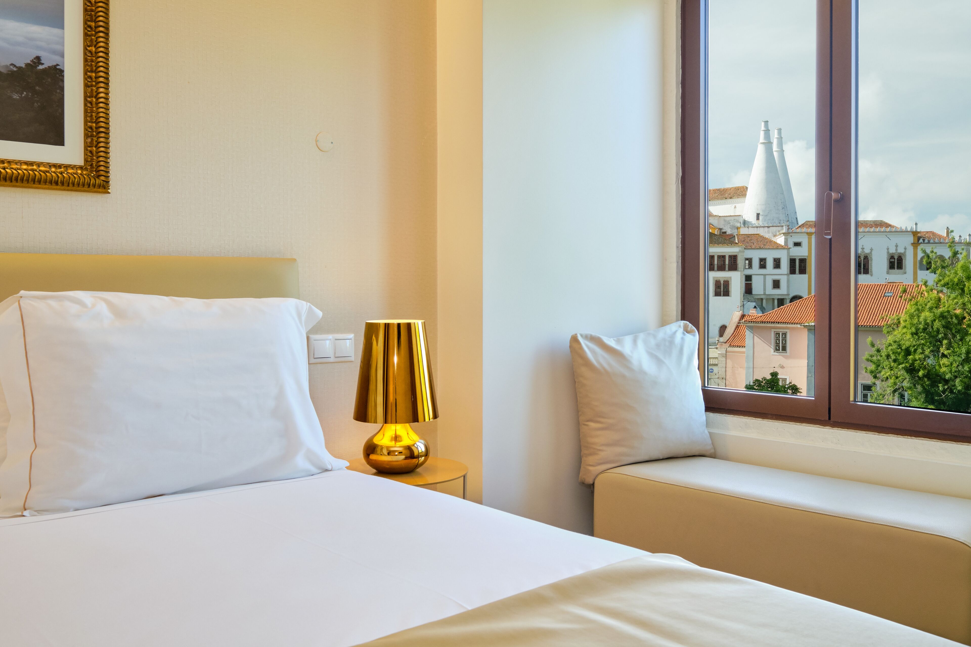 Habitación de hotel con cama, lámpara de noche, ventana con vistas a edificios y un cuadro colgado en la pared.