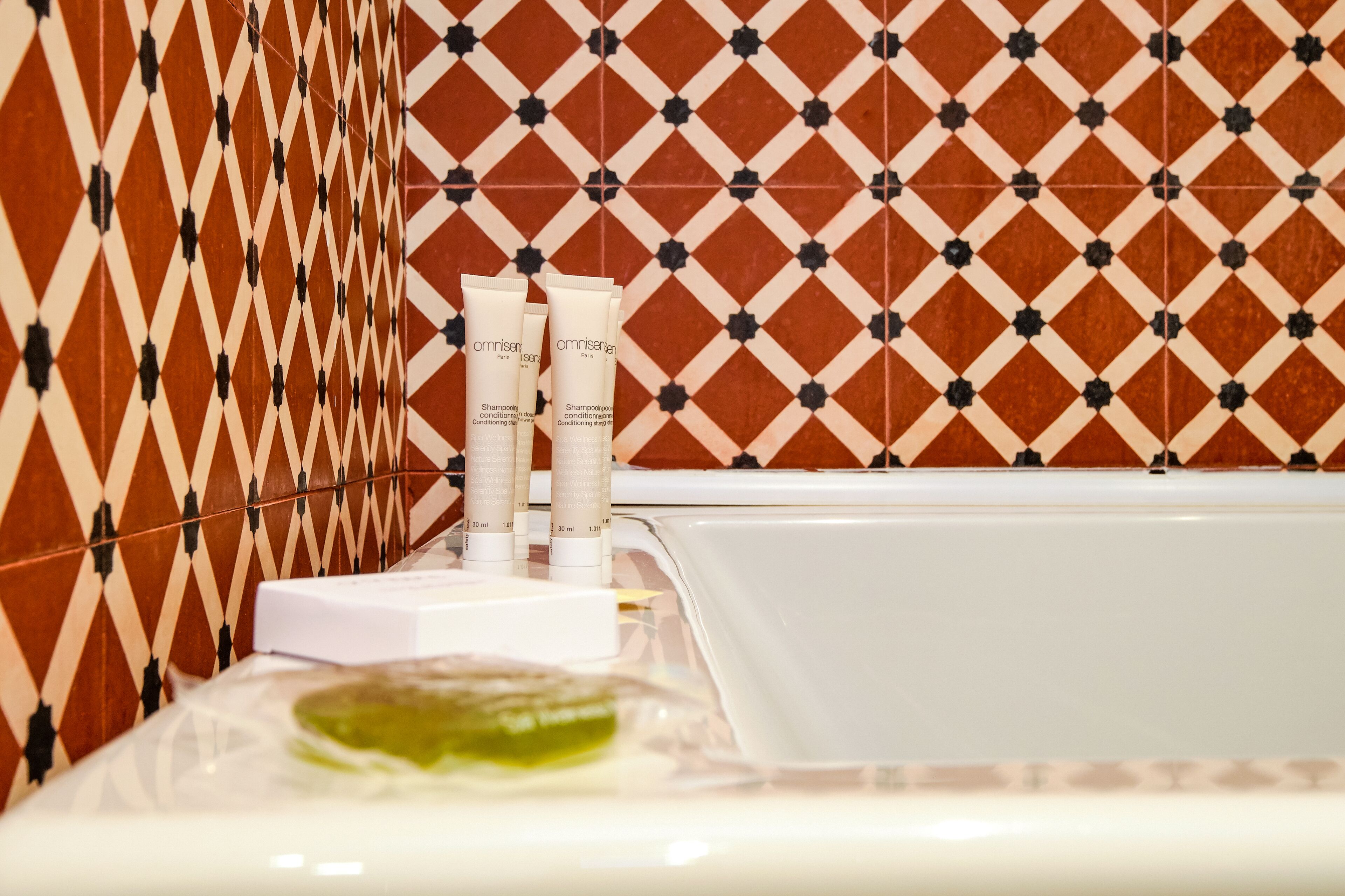 Un baño con pared de azulejos estampados, una bañera blanca y tres botellas de champú y acondicionador Omniscent.