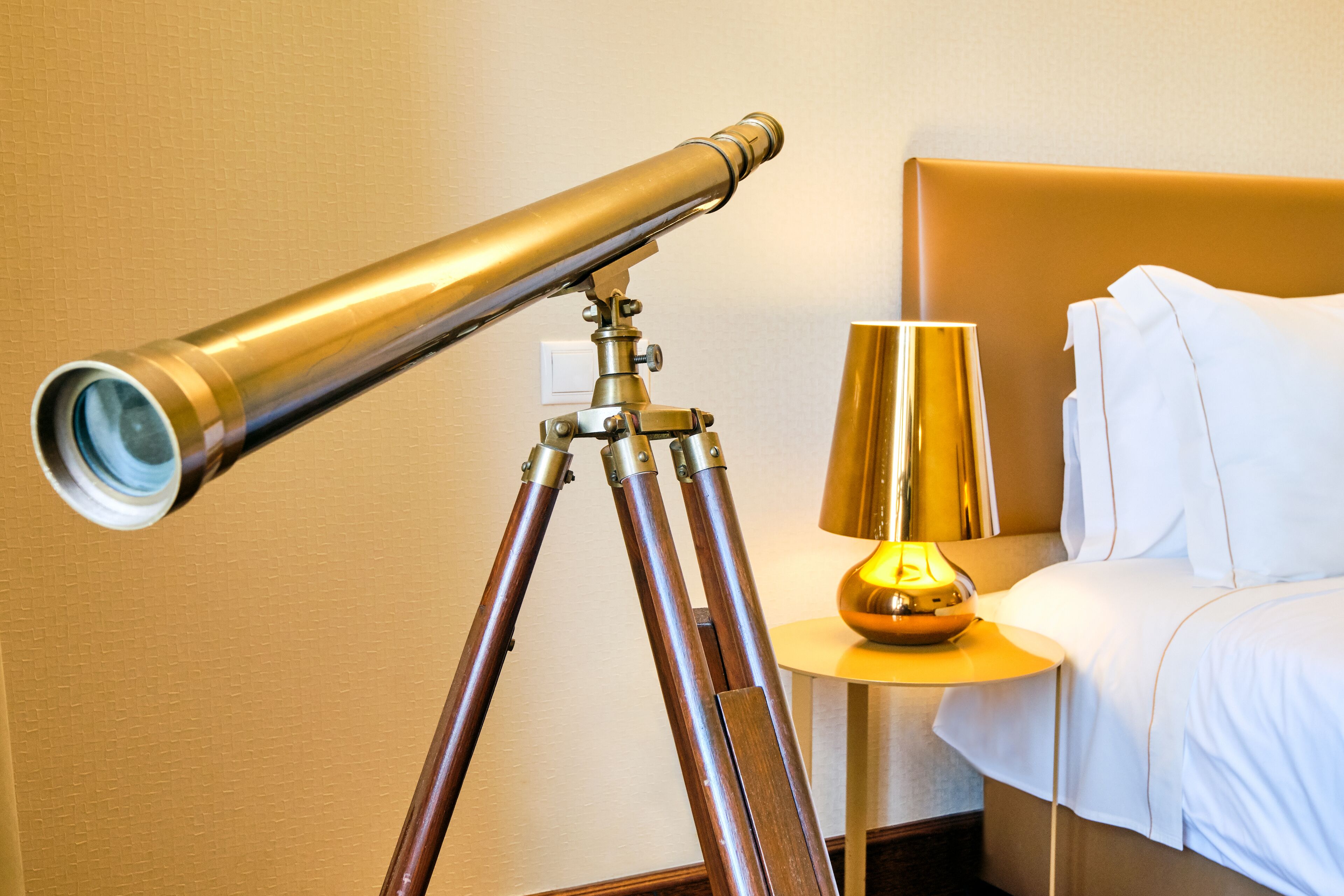 A telescope on a tripod, a lamp, and a bed in a hotel room.