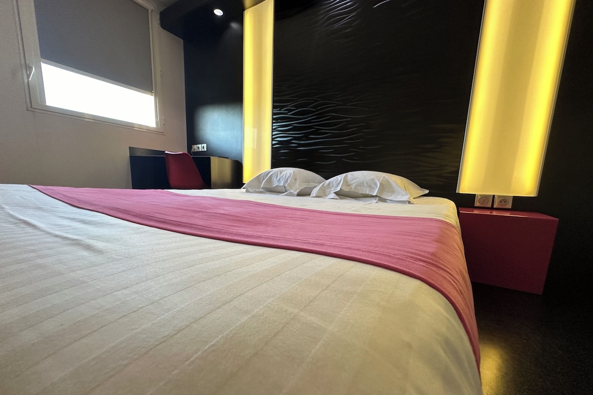 Habitación de hotel moderna con una cama grande, un escritorio y una silla. Cuenta con una ventana que proporciona luz natural y una pared con una textura distintiva.