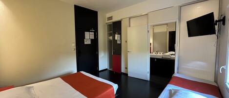 Una habitación de hotel con dos camas individuales, un baño con espejo y lavamanos, y un armario.