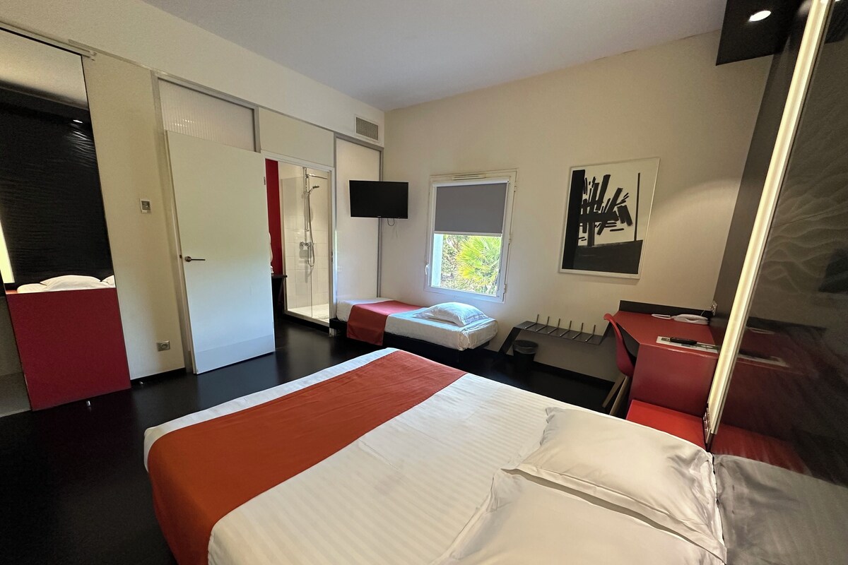 Habitación de hotel con una cama grande, zona de ducha, un pequeño escritorio y ventana con vistas a zonas verdes.