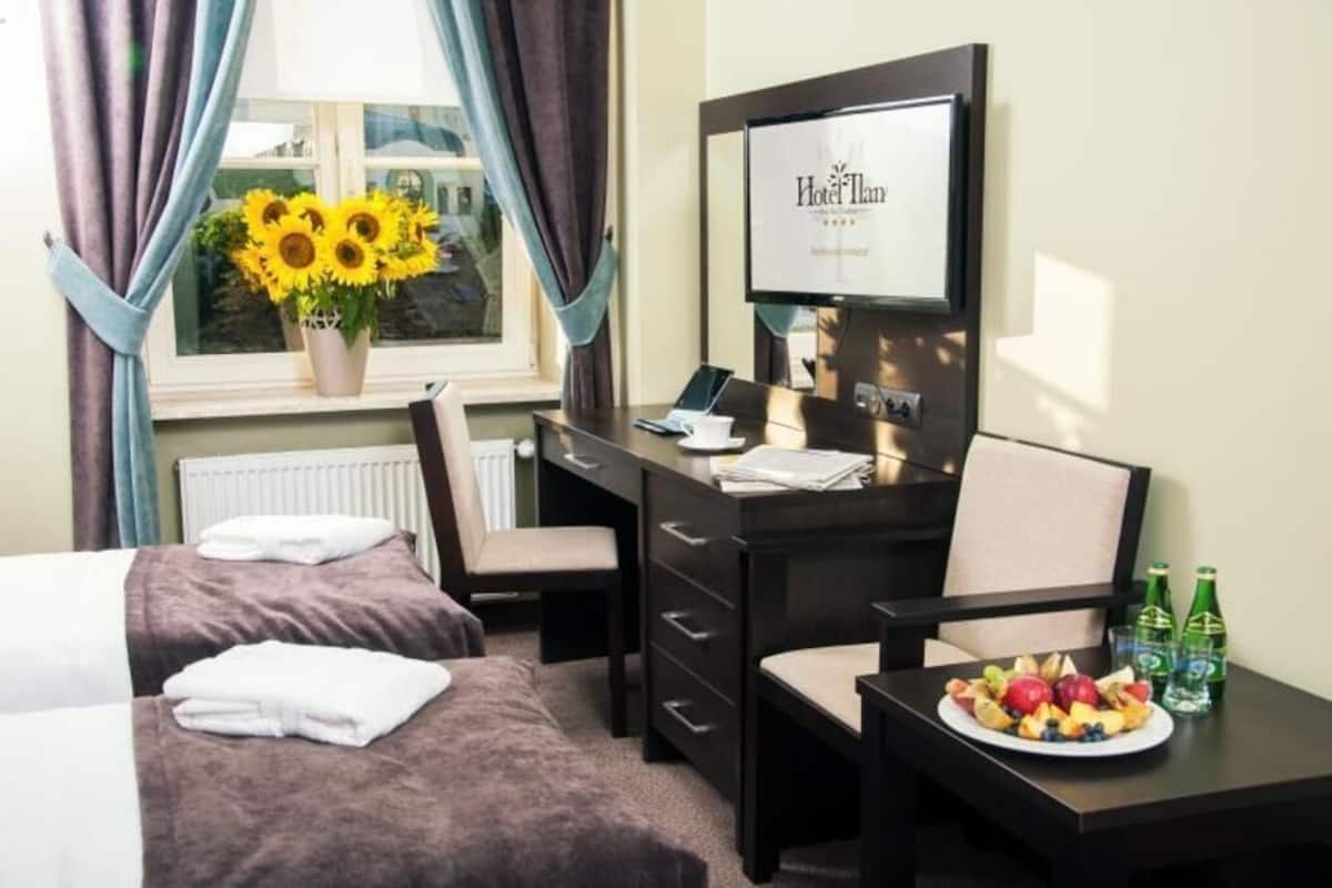 A hotel room with a bed, a desk with a computer, a TV, and a dining table with fruit and drinks.