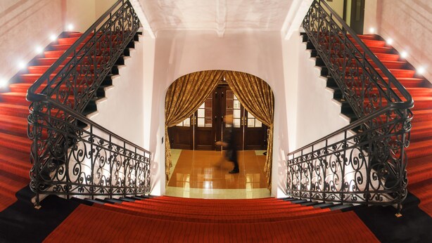 Eine prächtige Treppe mit aufwendigen schwarzen Geländern und roten Teppichstufen führt zu einem Raum mit einem großen, rundbogigen Durchgang und goldenen Vorhängen.