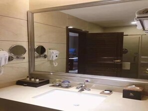 Un baño moderno con un espejo grande, un lavamanos, un espejo de mano, un secador de pelo y un dispensador de toallas.