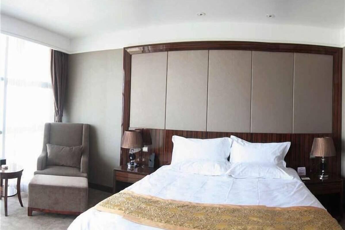 Ein Hotelzimmer mit einem großen Bett, einem Sessel, zwei Nachttischen mit Lampen und Blick ins Freie.