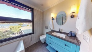 Une salle de bain avec un sol en parquet à chevrons, des toilettes, un lavabo avec un miroir et une fenêtre donnant sur un plan d’eau et de la verdure.