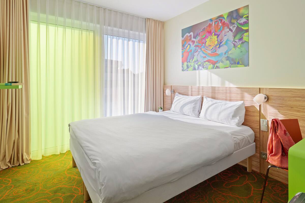 A hotel room with a large bed, a small table with a magazine, a lamp, and a colorful painting on the wall.