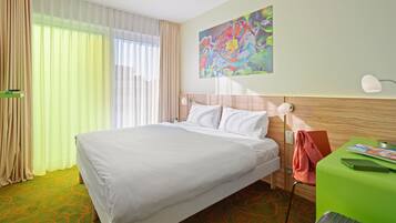 Ein Hotelzimmer mit einem großen Bett, einem kleinen Tisch mit einer Zeitschrift, einer Lampe und einem farbenfrohen Bild an der Wand.