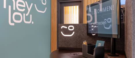 Un mostrador de recepción con un monitor que muestra 'WILLKOMMEN!' y una pared con el texto 'hey Lou'.