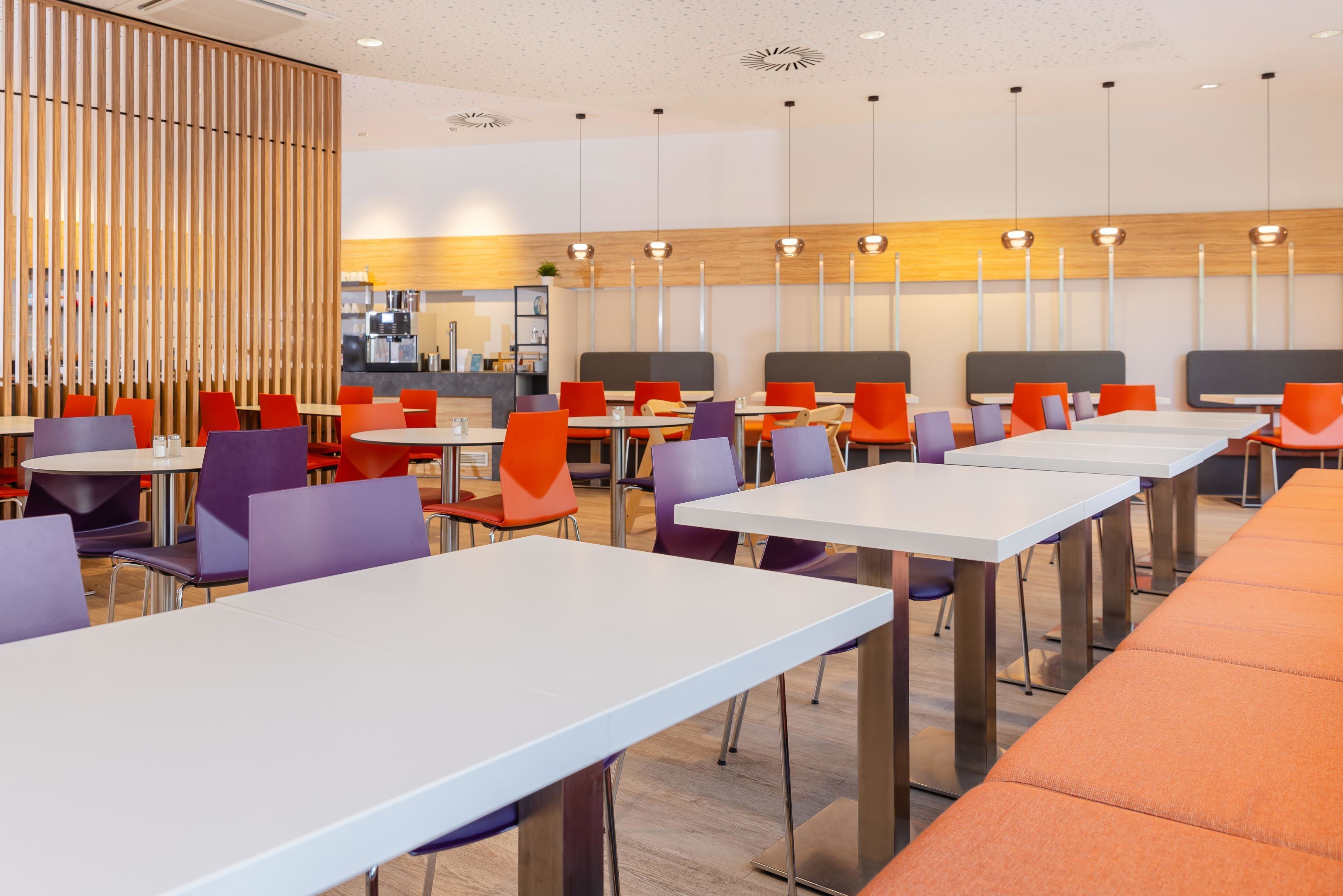 A modern dining area with multiple tables and chairs, a kitchen in the background, and a wooden partition.