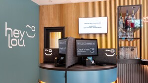 A reception desk with a sign that reads 'WILLKOMMEN!' and a digital screen displaying 'Check in here'. - Hey Lou Hotel Hildesheim (Hildesheim)
