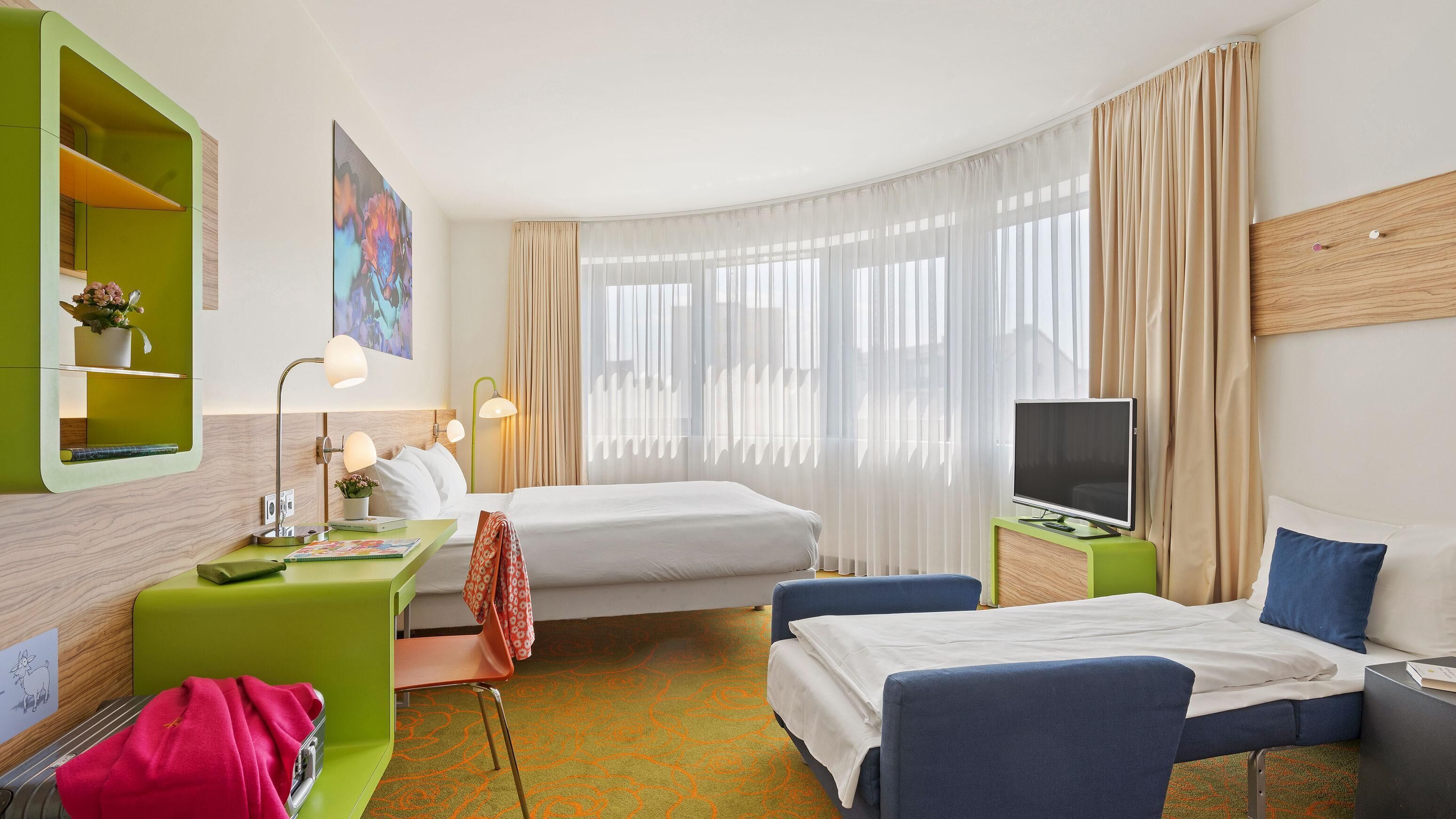 A hotel room with a bed, a desk with a chair, a TV, and a green seating area.