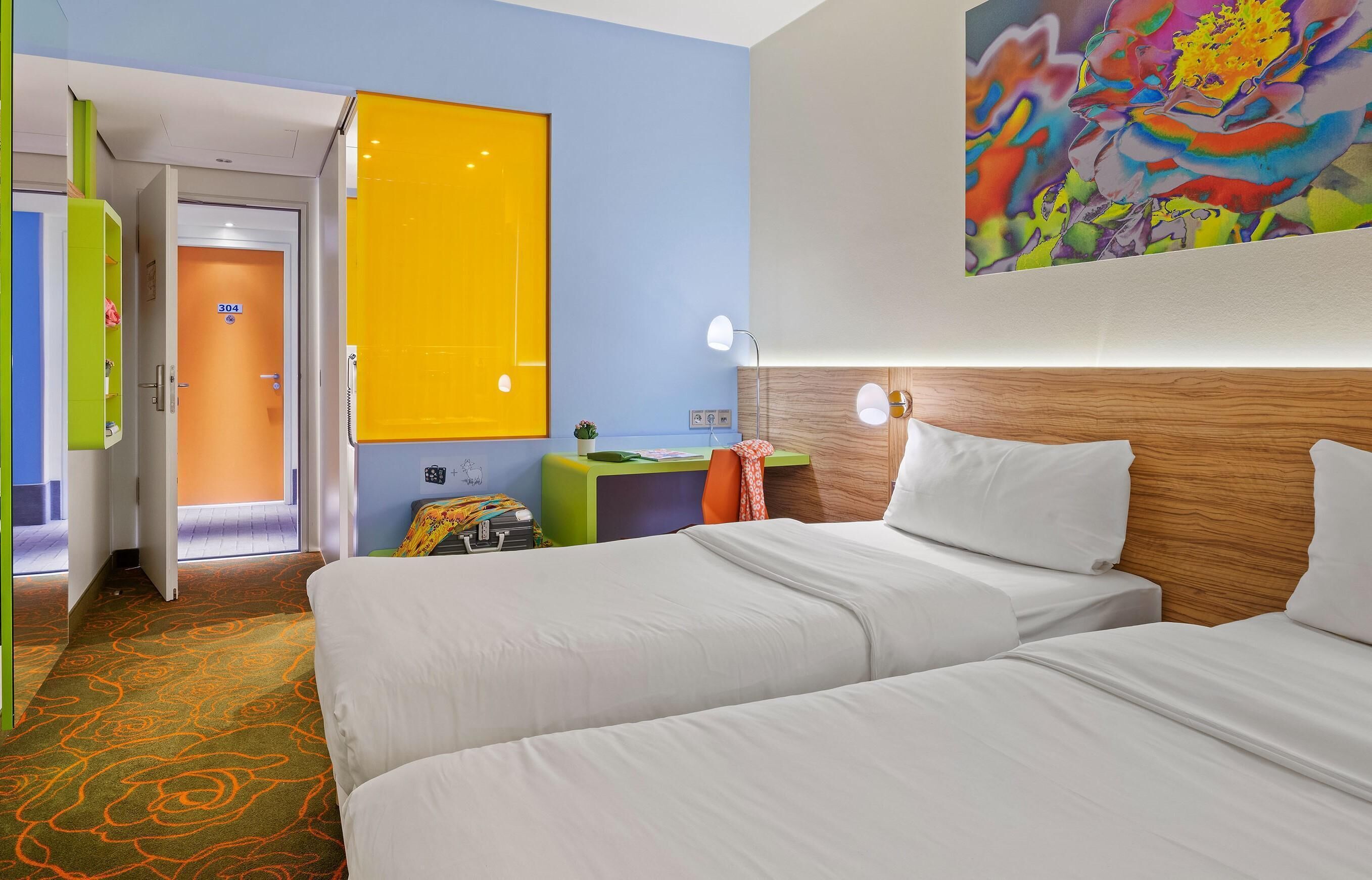 A hotel room with two beds, a wooden headboard, a colorful painting, a desk with a lamp, and a door with a number.