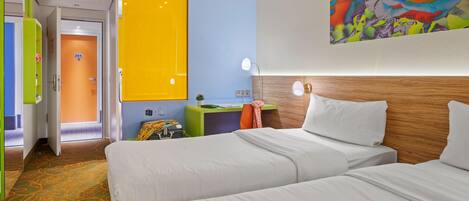 A hotel room with two beds, a wooden headboard, a colorful painting, a desk with a lamp, and a door with a number.