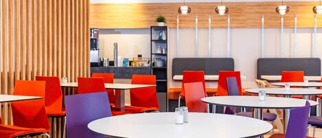 Un comedor moderno con mesas redondas y sillas de colores, una pared de acento de madera y un área de descanso con un banco gris.