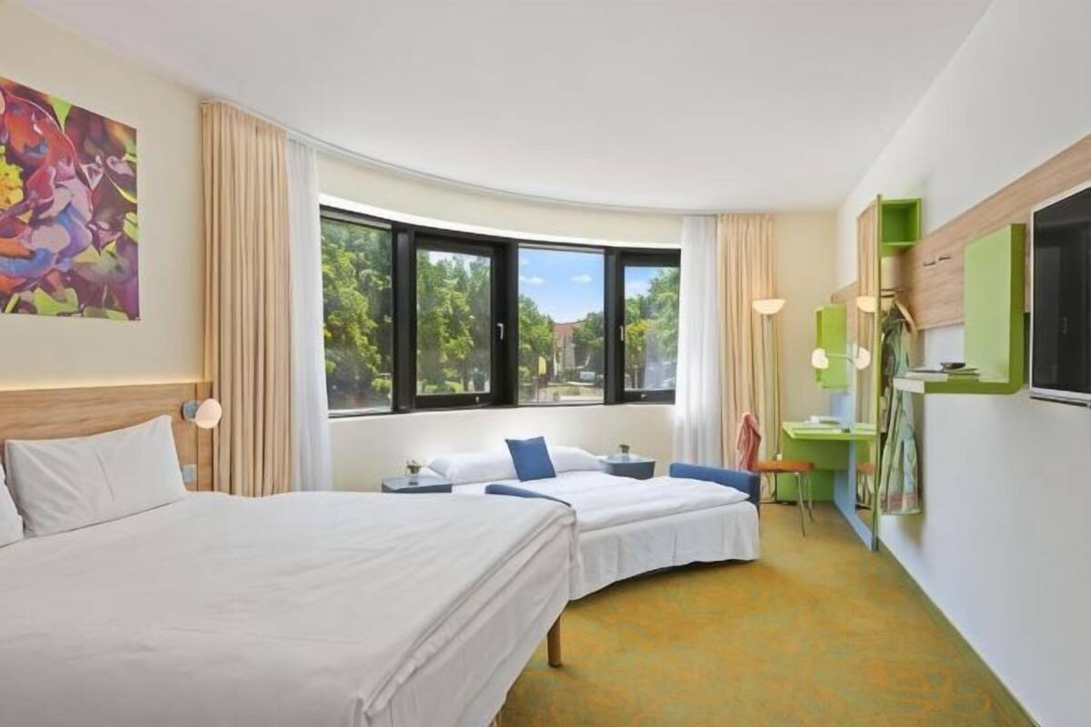 A hotel room with two beds, a desk, a chair, a window with a view, and a painting on the wall.