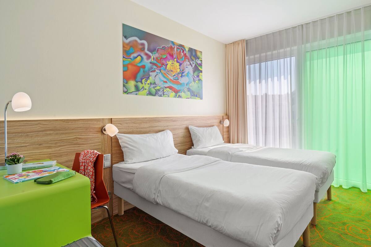A hotel room with two beds, a desk with a lamp, a painting on the wall, and a large window with curtains.