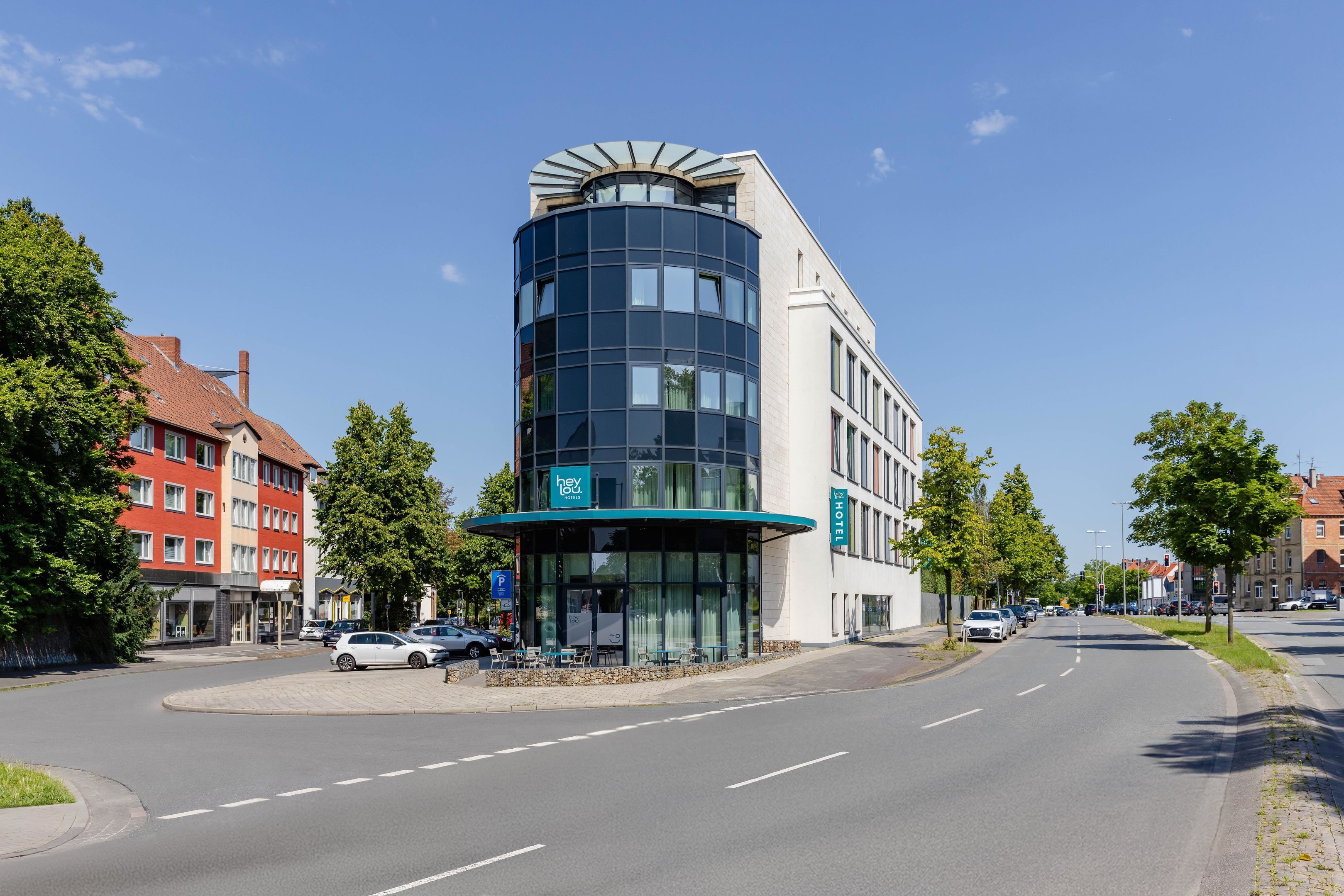 Ett modernt hotell med en böjd glasfasad, en parkeringsplats och utsikt över gatan med träd och andra byggnader.