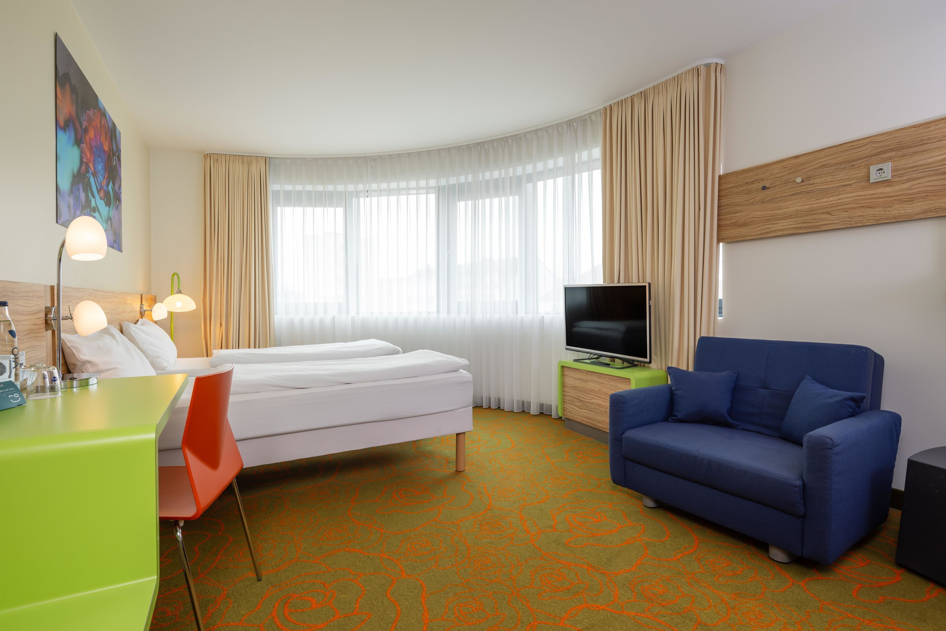 A hotel room with a bed, a TV, a blue sofa, a desk with a lamp, and a large window with curtains.