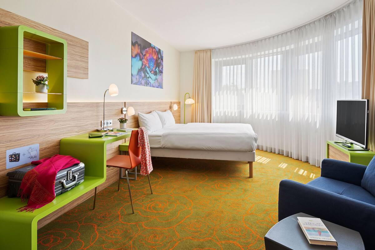 A hotel room with a bed, a desk, a chair, a sofa, a TV, and a painting on the wall.