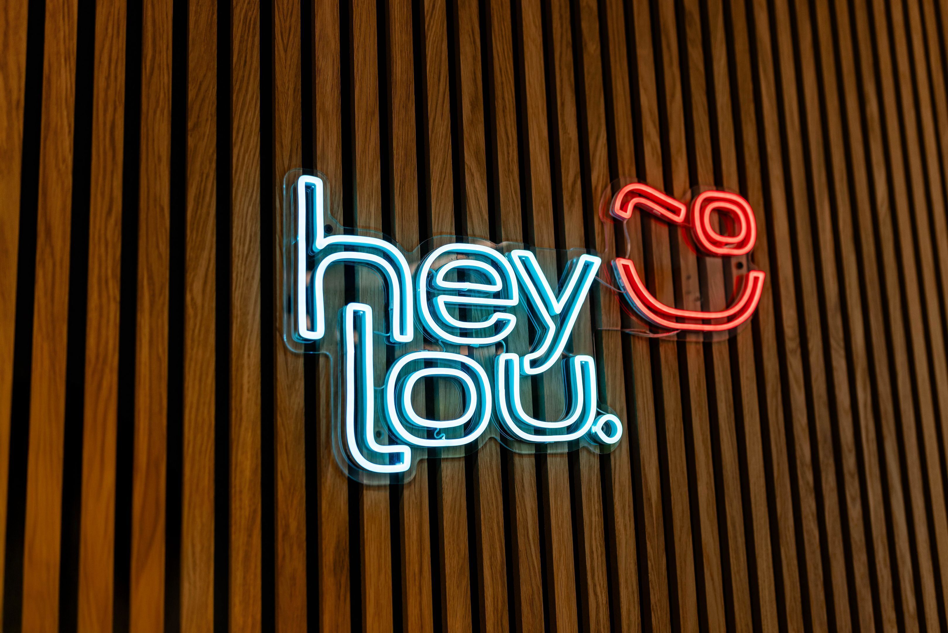 A neon sign with the text 'hey Lou.' on a wooden slatted background.