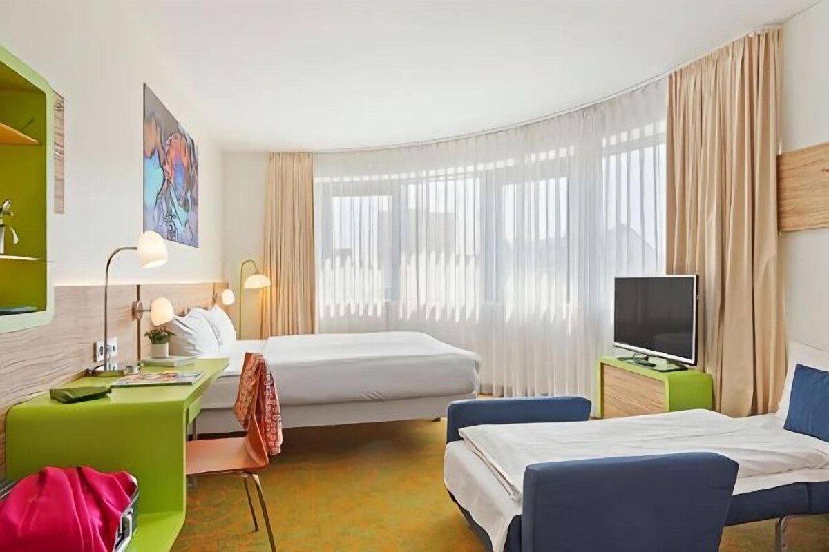 A hotel room with two beds, a desk, a chair, a TV, and a large window with sheer curtains.