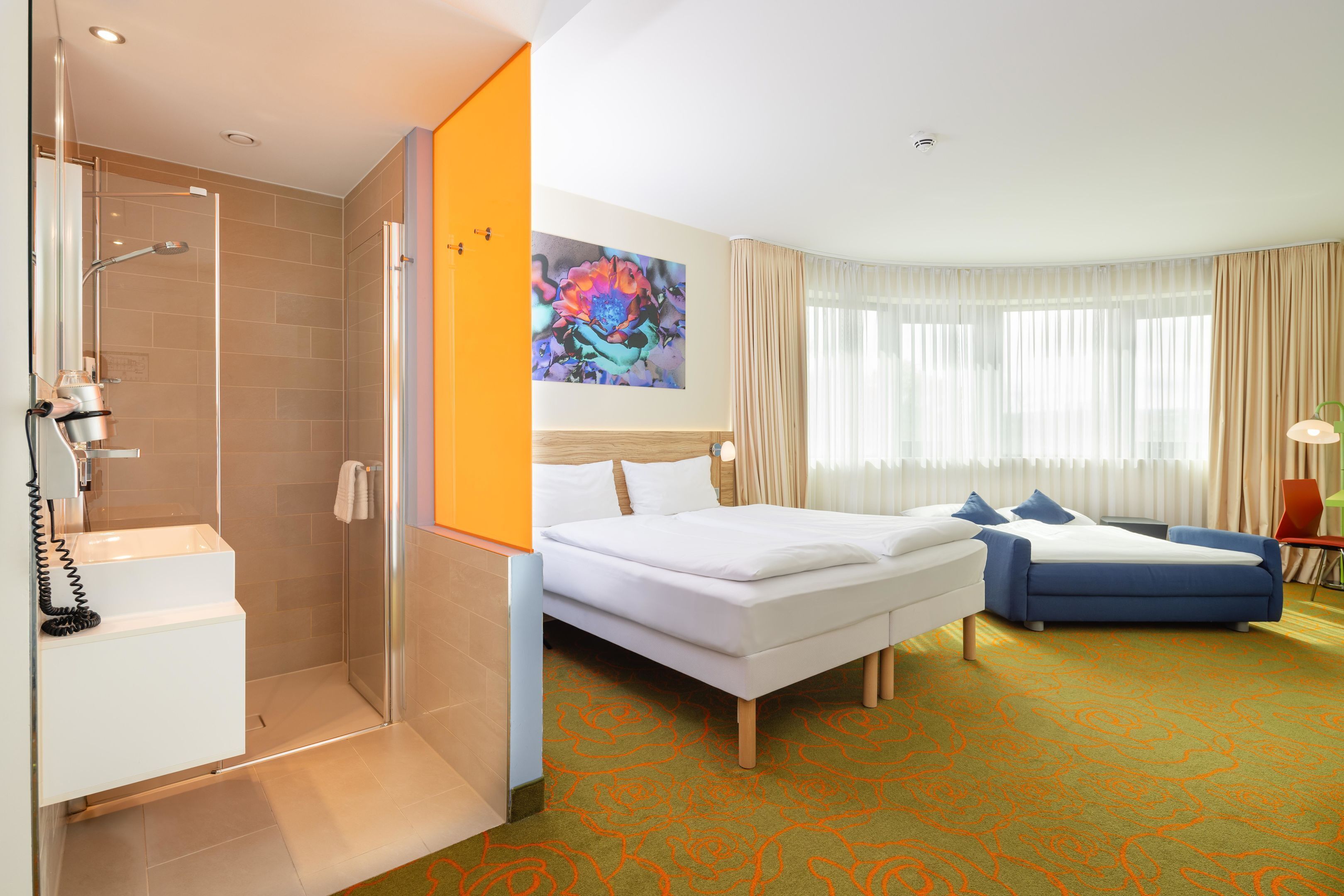 A hotel room with a shower, a bed, a bedside table, a lamp, and a window with curtains.