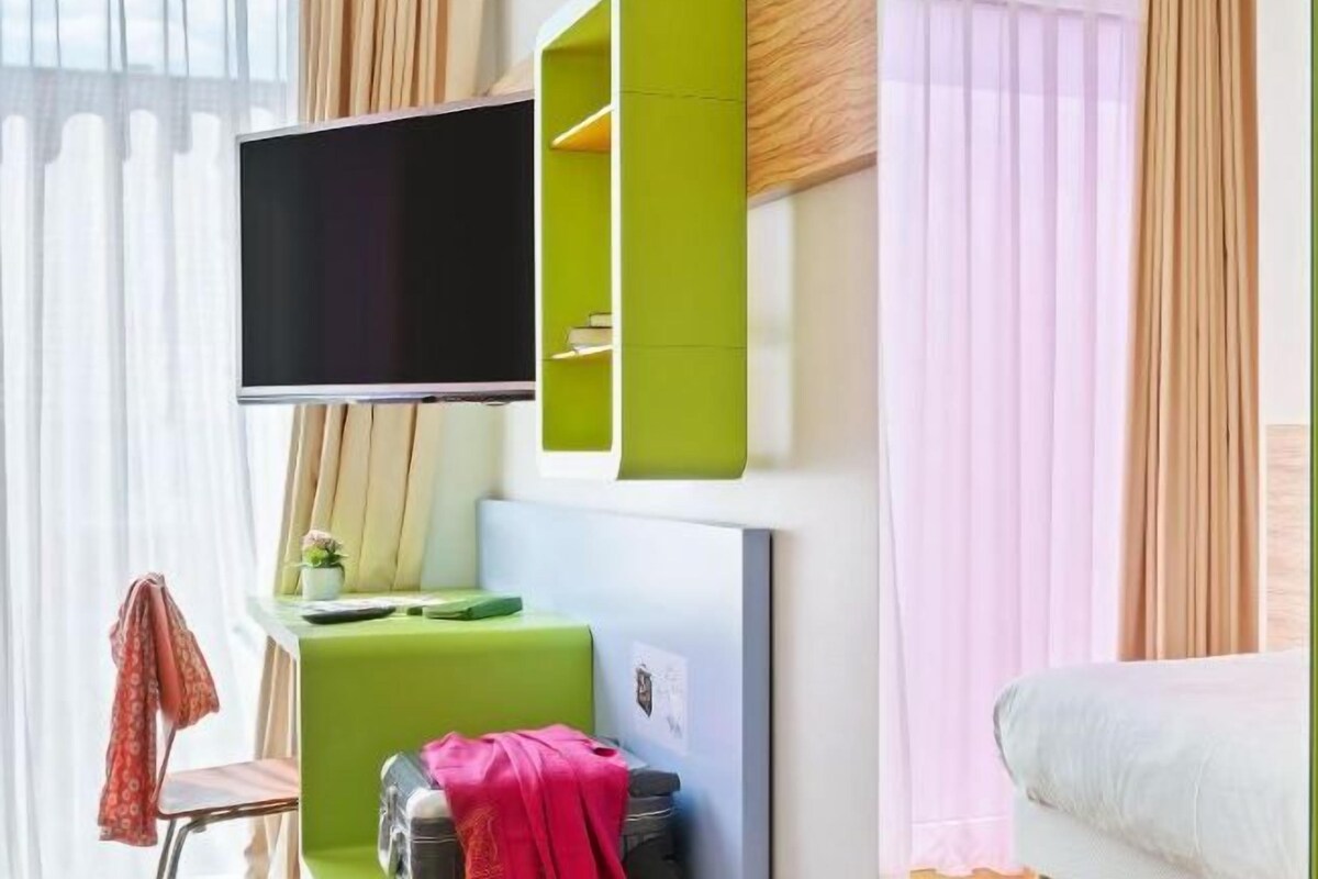 A modern room with a flat-screen TV mounted on the wall, a green desk with a chair, a bed with a white comforter, and a window with sheer curtains.