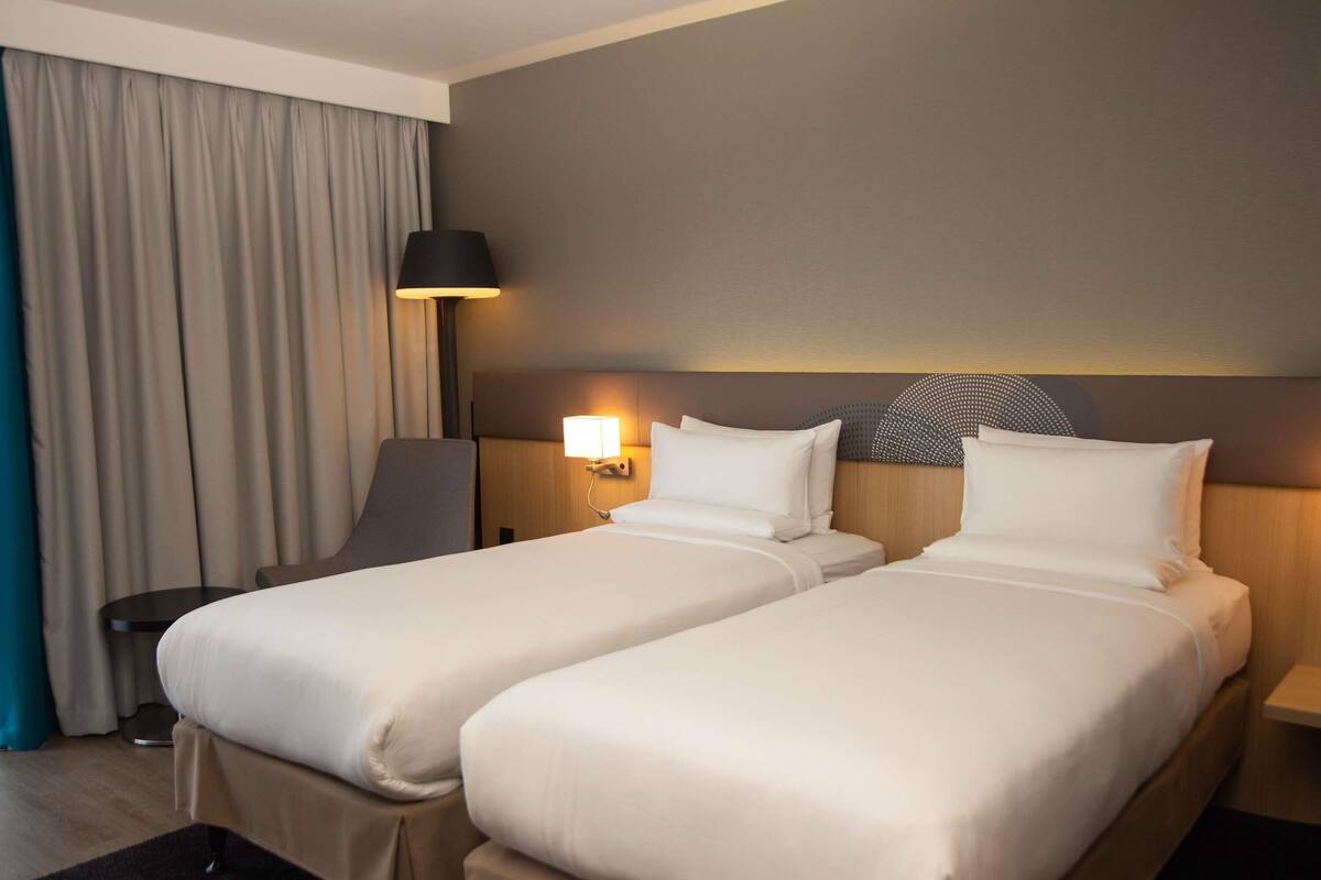 A hotel room with two single beds, a desk, a chair, a lamp, and a large window with curtains.